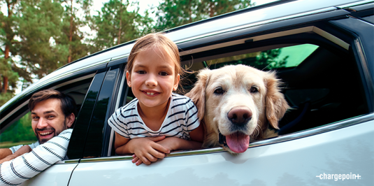 A child, a dog and a parent share joyous smiles as they peer out of a car window.